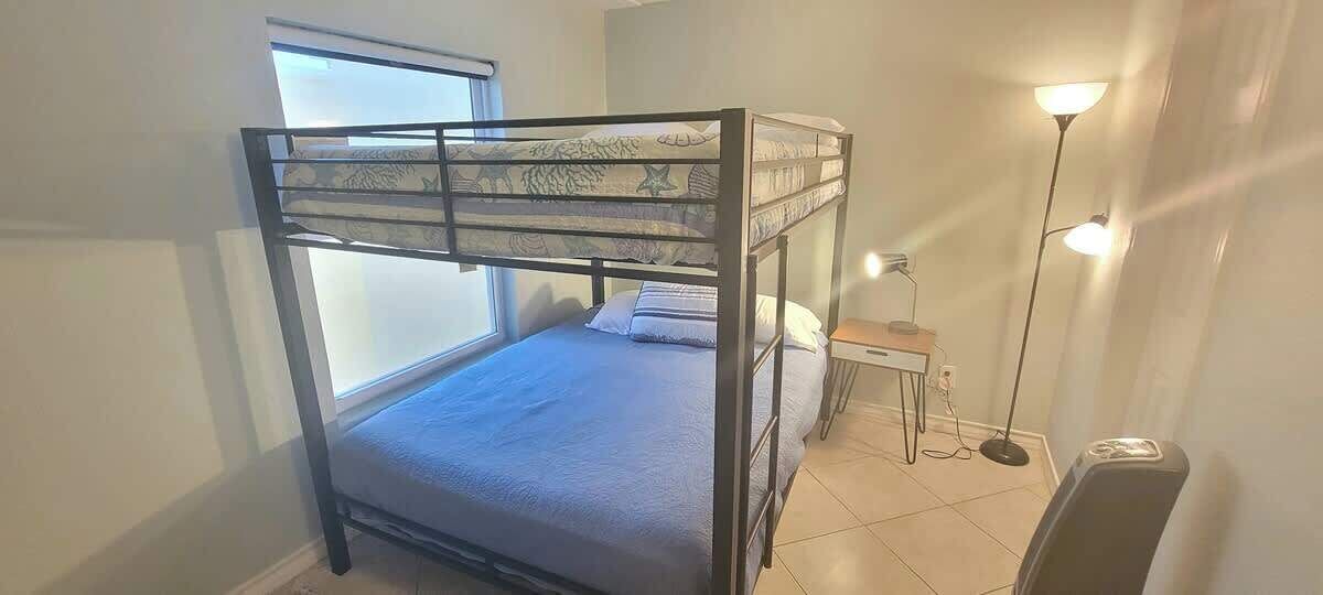 A bunk bed with a patterned headboard, a bedside table with a lamp, and a window with blinds.