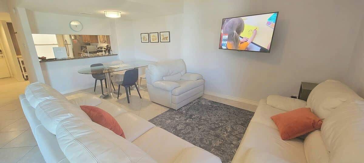 Un salon avec un canapé blanc, une table à manger avec des chaises, un téléviseur fixé au mur et, au fond, un coin cuisine.