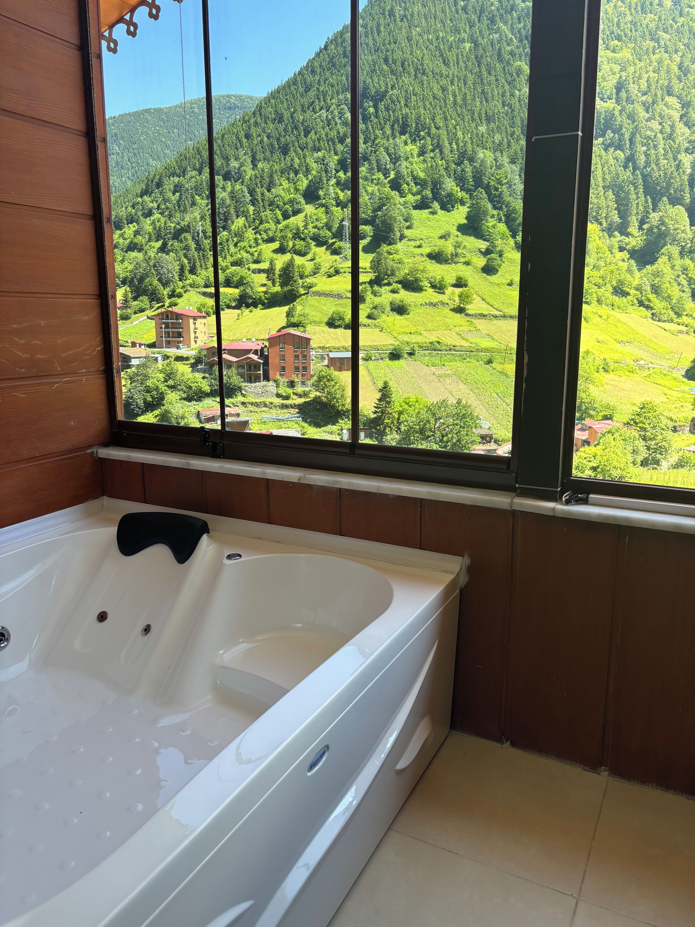 A bathroom with a large bathtub, wooden walls, and a window offering a view of a green valley with buildings.
