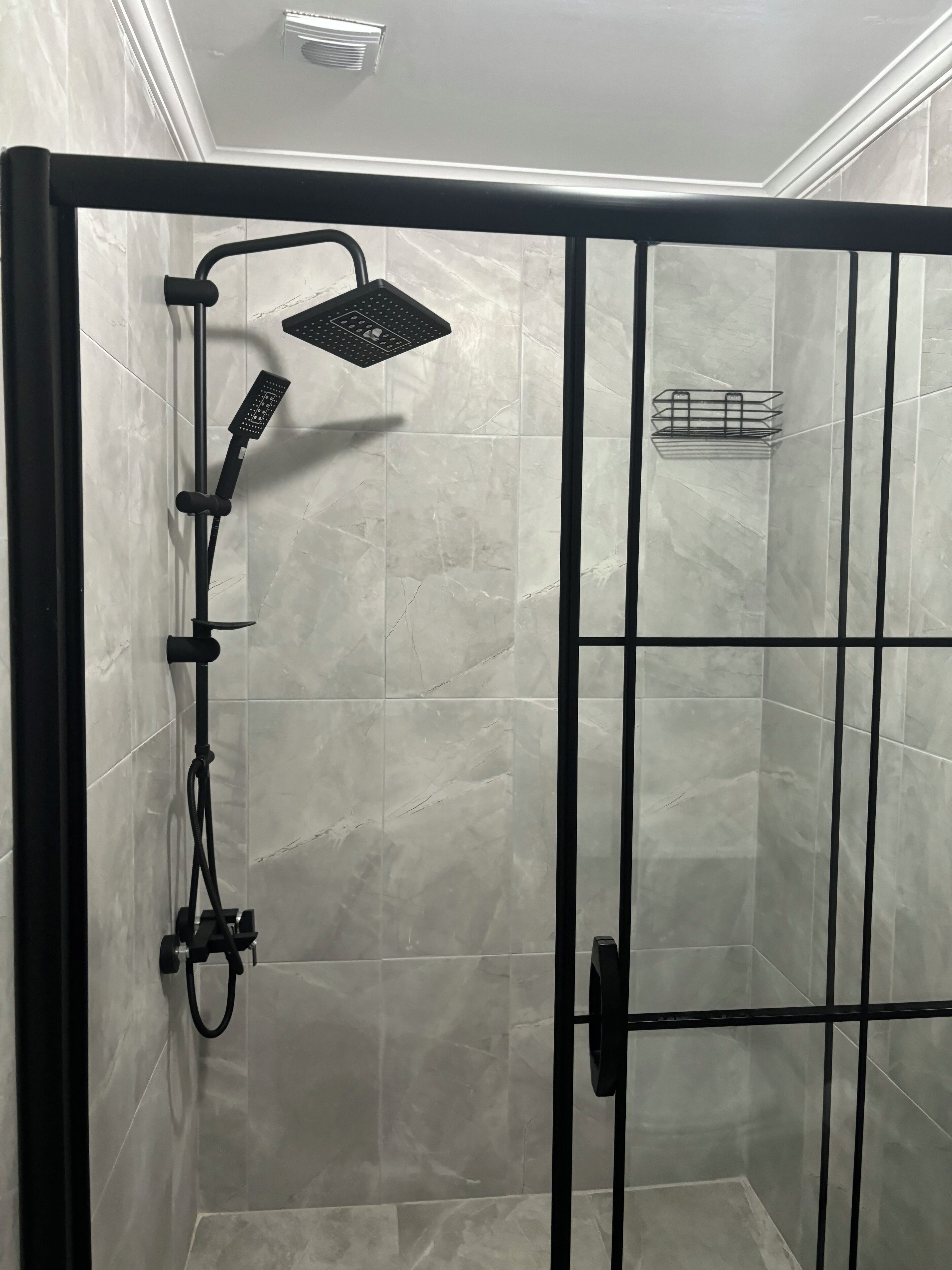 A walk-in shower with a black frame, a handheld showerhead, a wall-mounted control panel, and a small shelf with toiletries.