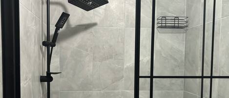 A walk-in shower with a black frame, a handheld showerhead, a wall-mounted control panel, and a small shelf with toiletries.