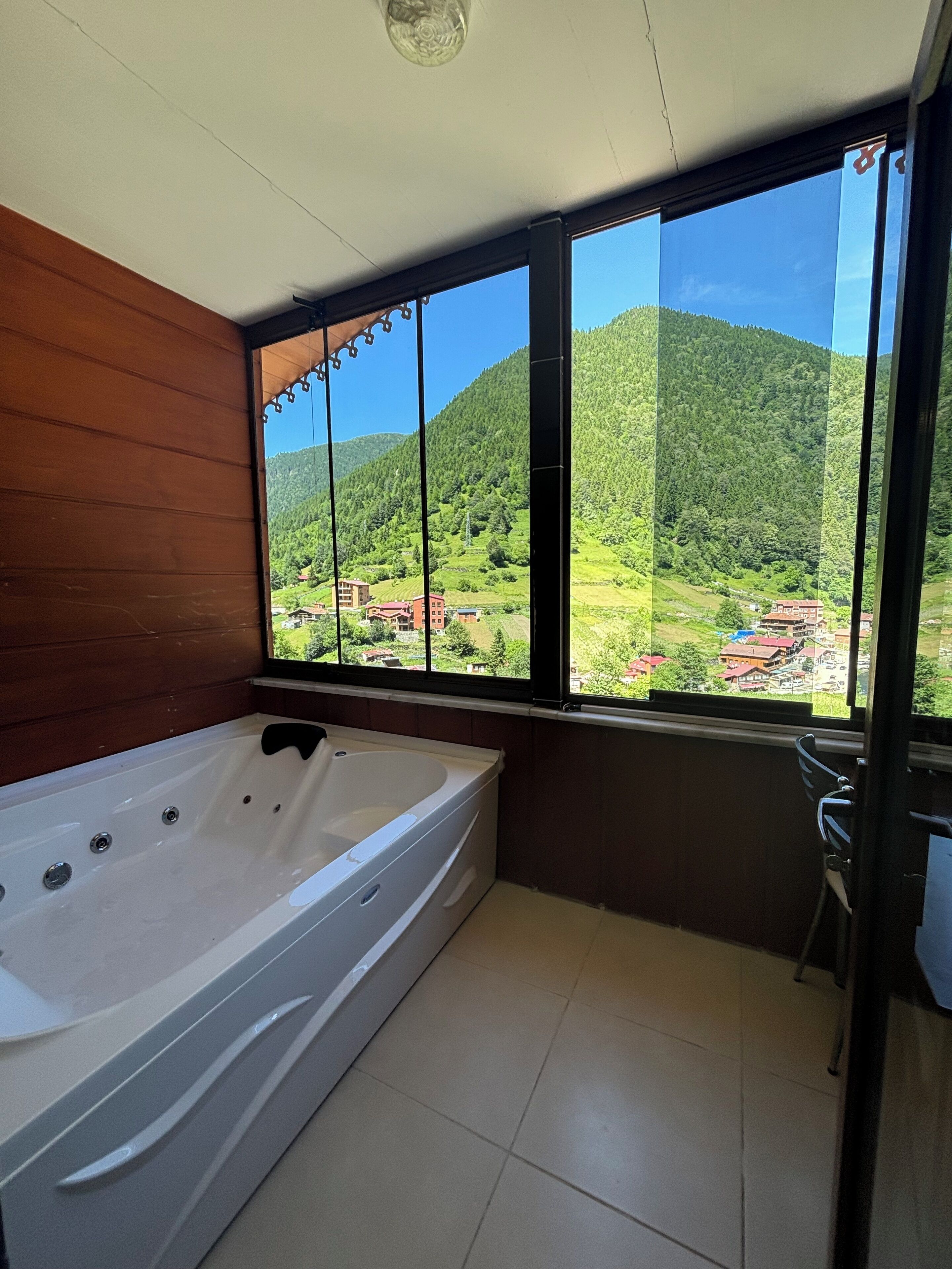 A bathroom with a large bathtub, a window with a view of a green valley and buildings, and a wooden wall.