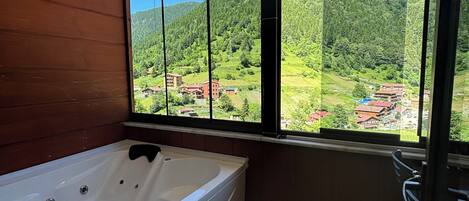 A bathroom with a large bathtub, a window with a view of a green valley and buildings, and a wooden wall.