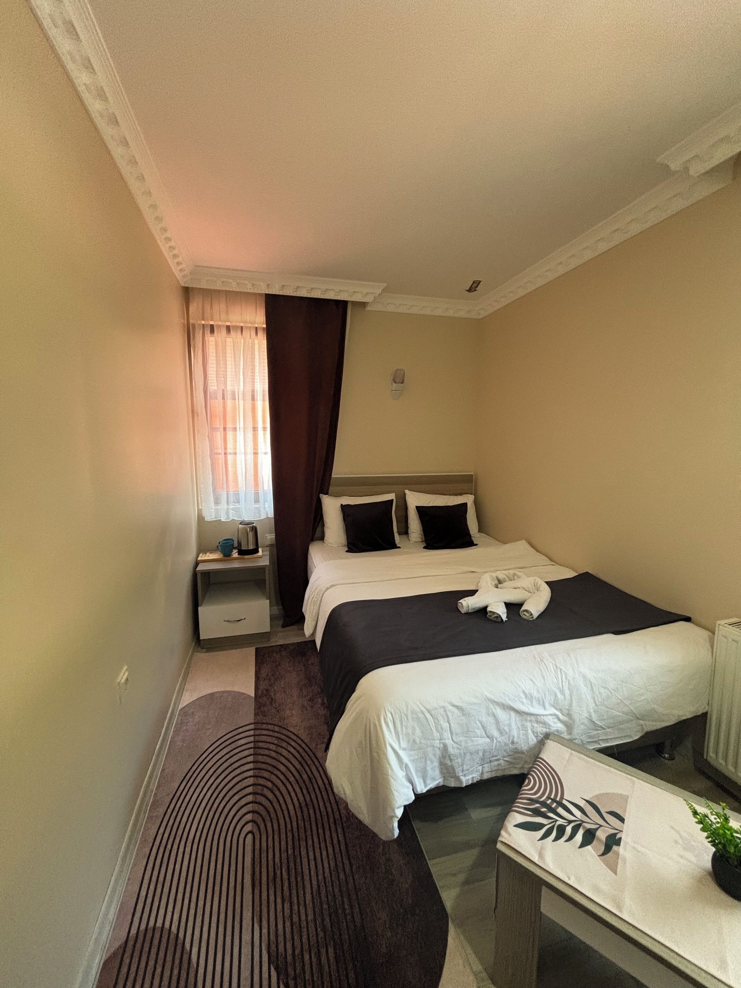 A hotel room with a bed, a bedside table, a nightstand, a window with curtains, and a radiator.