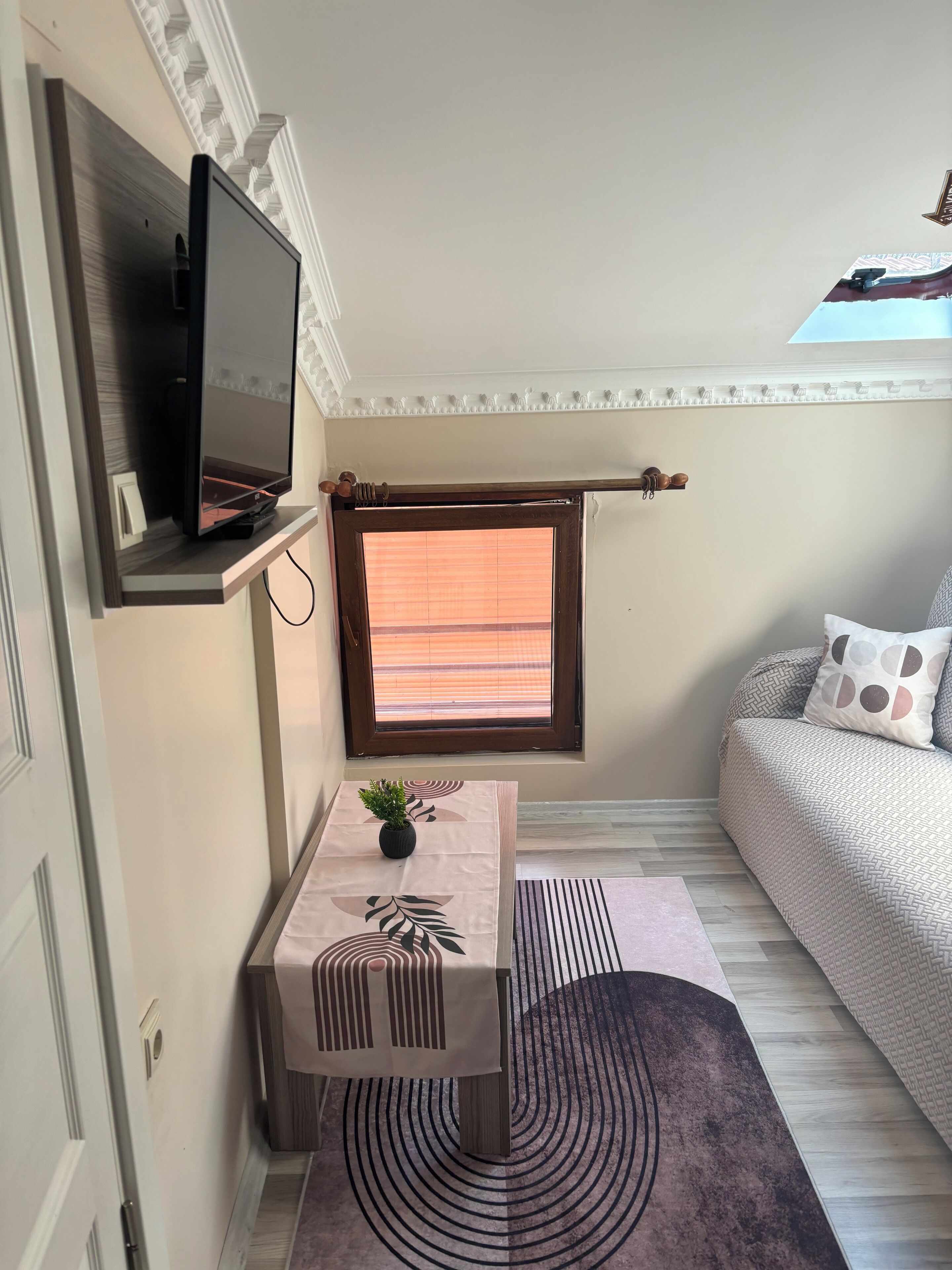 Una habitación moderna con una televisión de pantalla plana montada en la pared, una pequeña mesa con una planta, una ventana con persianas, un sofá con cojines estampados y un tragaluz.