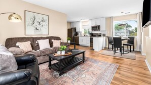 A modern living room with a sofa, coffee table, and a rug. A kitchen with white cabinets and a dining area with chairs is visible in the background.