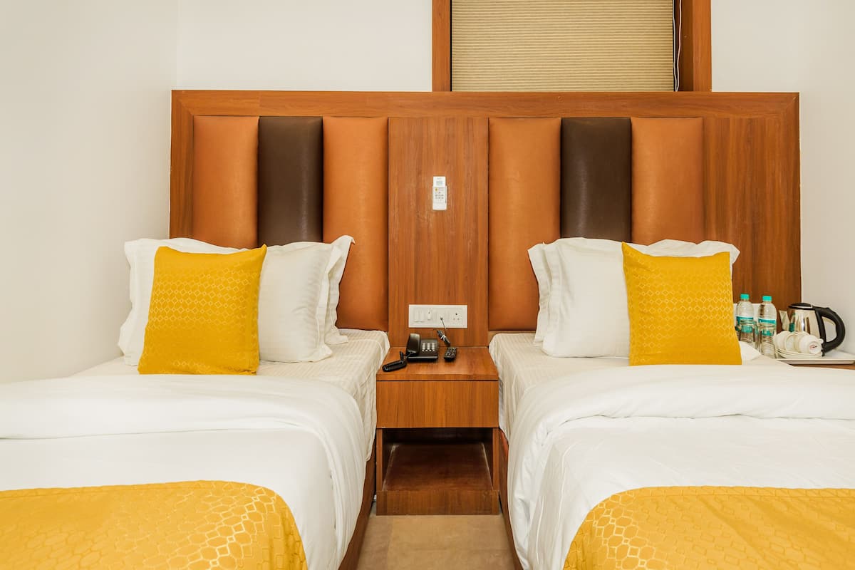 A hotel room with two beds, each with a wooden headboard and a yellow patterned bedspread. There is a nightstand between the beds with a phone, a kettle, and some water bottles.