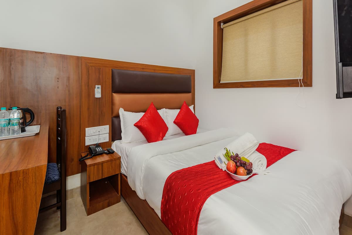 A hotel room with a wooden headboard, a bedside table with a phone and a kettle, a red throw blanket, and a fruit bowl on the bed.