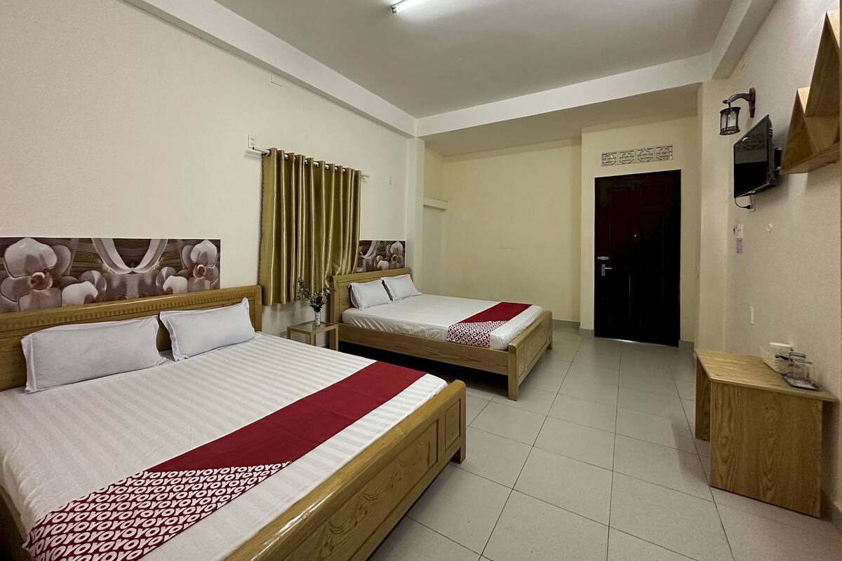 A hotel room with two beds, a wooden headboard, a small bedside table, a TV mounted on the wall, and a door leading to another room.