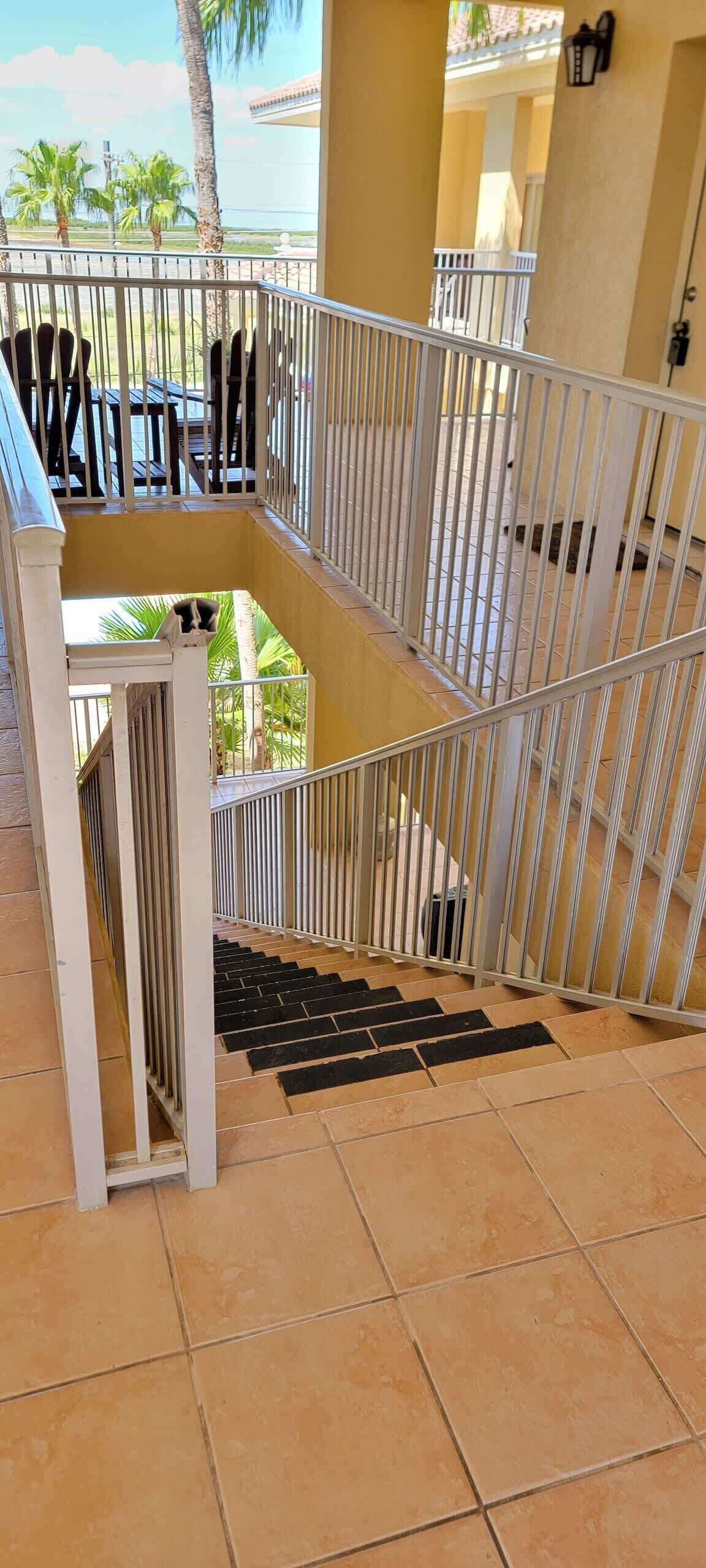 A multi-level outdoor staircase with metal railings, surrounded by a tiled floor and a view of palm trees and a building in the background.
