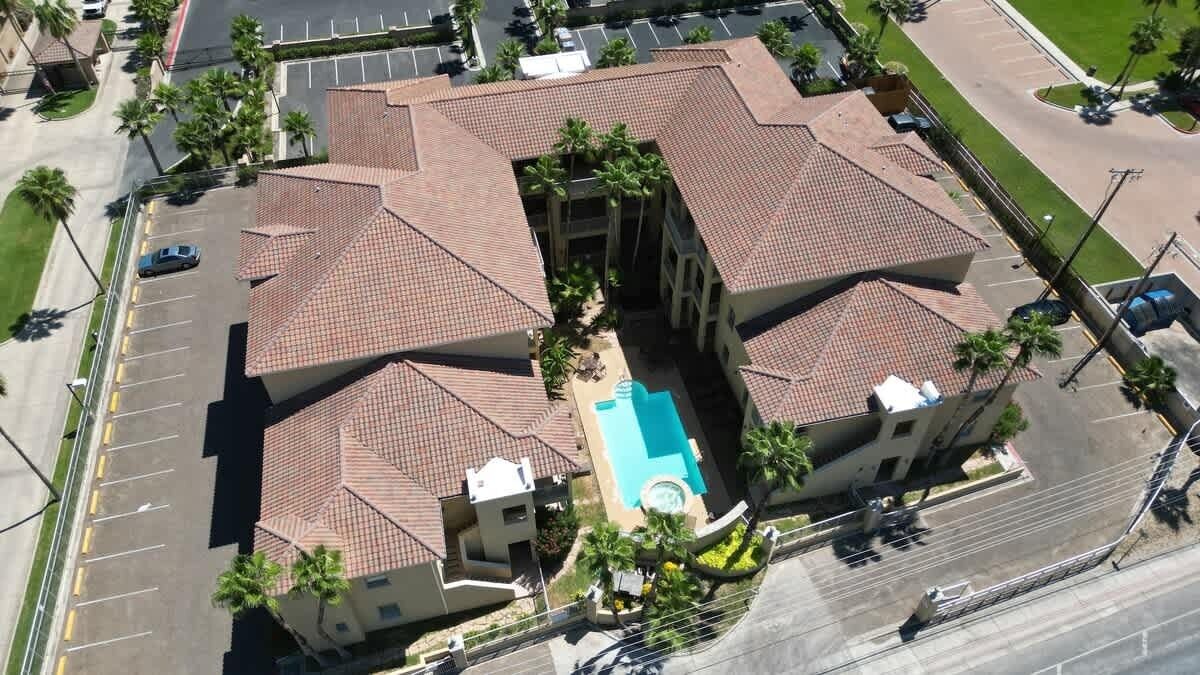 An aerial view of a building with a swimming pool, surrounded by palm trees and a parking lot.