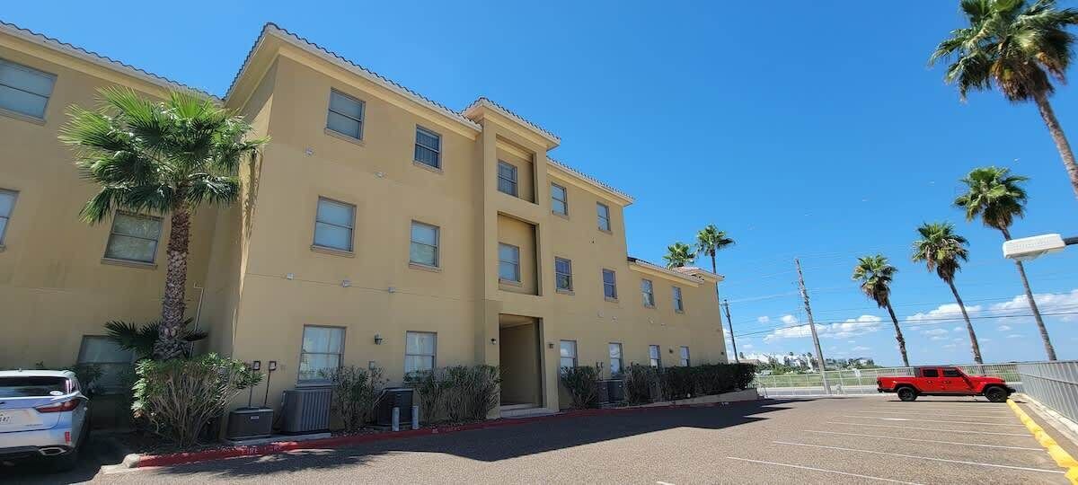 Un hôtel à plusieurs étages doté d’un parking, de palmiers et d’un véhicule rouge.