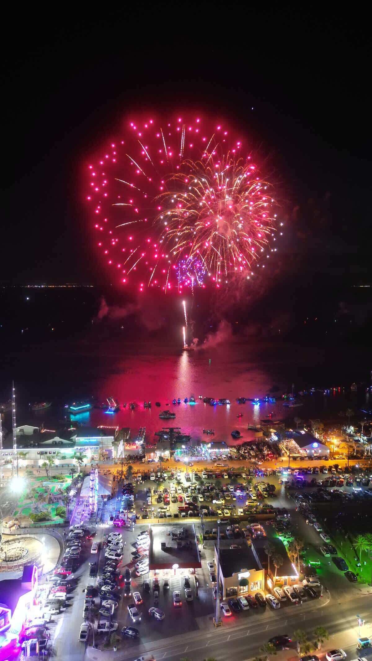 A nighttime view of a waterfront area with fireworks display, boats, and a parking lot.