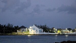 Un bâtiment situé sur la côte, avec une façade blanche et des fenêtres bleues, se dressant près d’une étendue d’eau, tandis qu’un croissant de lune brille dans le ciel.
