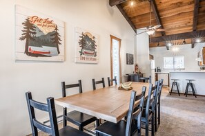 A dining area with a wooden table and chairs, a kitchen in the background, and two wall art pieces. - Private Lodge, Vaulted Ceilings, Gas Grill, Washer/Dryer, Walk to The Village (Mammoth Lakes)