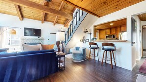 Un salon moderne avec un canapé bleu, une cheminée, un escalier et un coin cuisine doté de meubles en bois et de tabourets de bar.