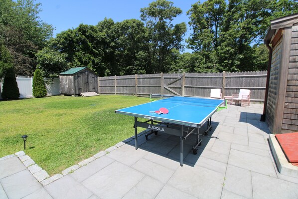 Une table de ping-pong dans un jardin, avec une clôture en bois et des arbres en arrière-plan.