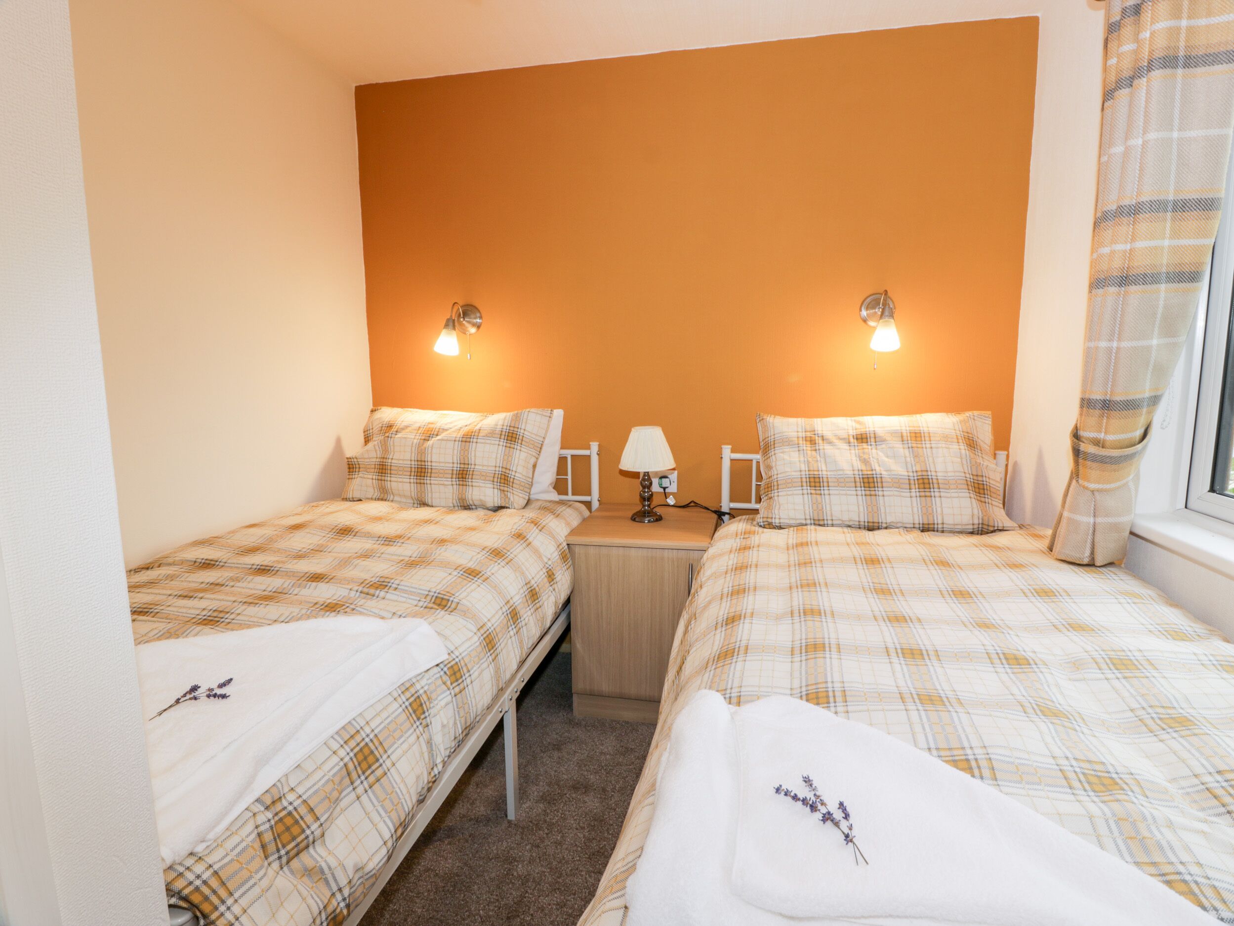 A twin room with two beds, each with a plaid duvet, a bedside table with a lamp, and a wall-mounted light fixture.