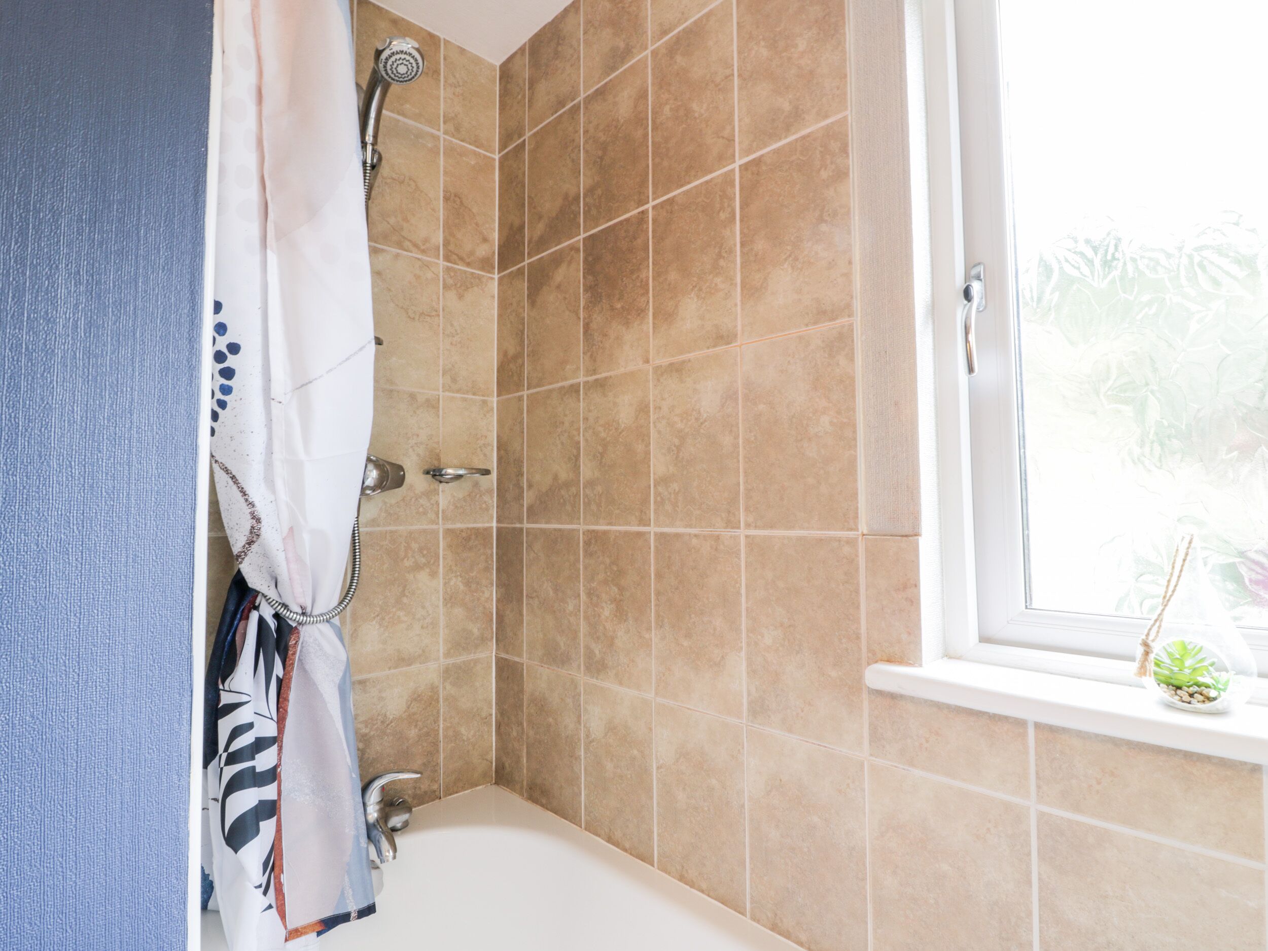 Une salle de bain avec des carreaux de couleur beige, une douche, une fenêtre donnant sur un espace verdoyant et une serviette accrochée à la porte.