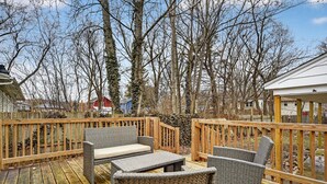 A wooden deck with wicker furniture, including a sofa and chairs, surrounded by trees and a red building in the background. - Southern Comfort Cottage in the City (Lansing)