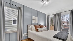 A bedroom with a bed, curtains, a window with blinds, a wall-mounted artwork, and a view of the outside. - Southern Comfort Cottage in the City (Lansing)