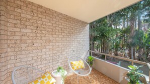 A balcony with a brick wall, two white wire chairs with yellow cushions, a potted plant, and a view of palm trees. - Breezy, Stylish, Getaway (Tuncurry)