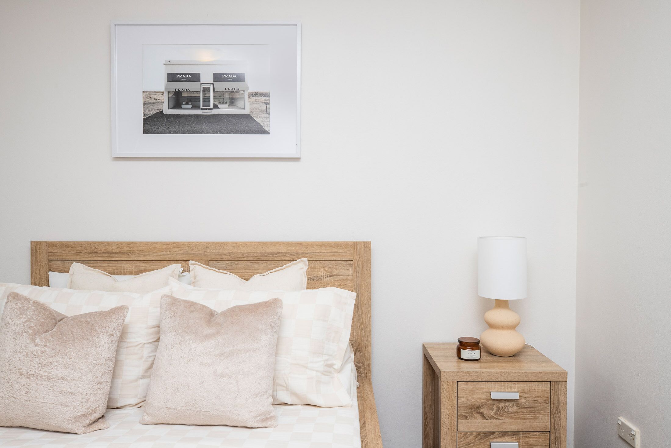Een netjes opgemaakt bed met een houten hoofdbord, een nachtkastje met een lamp en een klein vaasje, en een lijst met een afbeelding van een gebouw aan de muur.