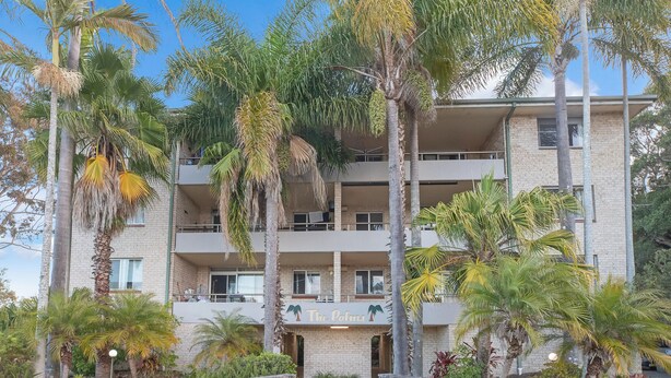 A multi-story building with a sign reading "The Palms." It features a brick facade, multiple balconies, and lush tropical landscaping. - Breezy, Stylish, Getaway (Tuncurry)
