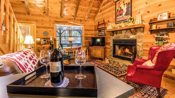 A cozy log cabin interior with a stone fireplace, a red armchair, and a dining area with a tray of wine and glasses. - Black Bear Bungalow - Lovely Chalet in the Woods (Clarkesville)