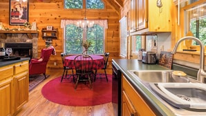 A cozy cabin interior with a dining area, wooden walls, a stone fireplace, and a large window. - Black Bear Bungalow - Lovely Chalet in the Woods (Clarkesville)