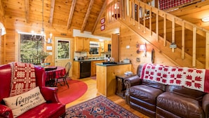 A cozy cabin interior with a dining area, a living room with a leather couch, a kitchen, and a wooden staircase. - Black Bear Bungalow - Lovely Chalet in the Woods (Clarkesville)