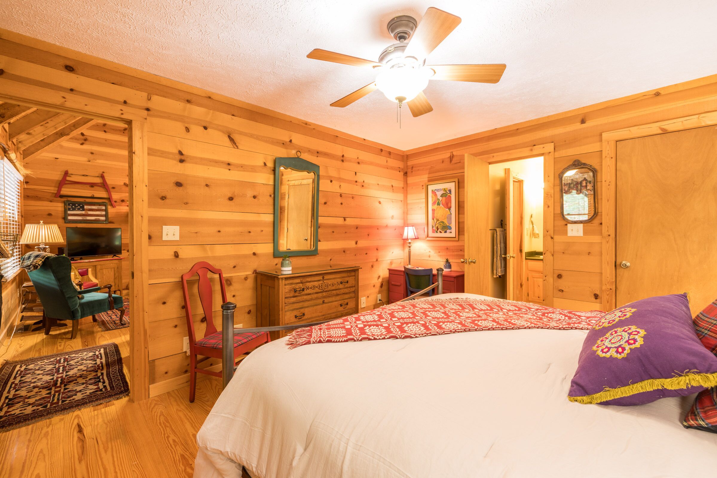 A wooden cabin bedroom with a bed, a chair, a dresser, a mirror, and a ceiling fan.