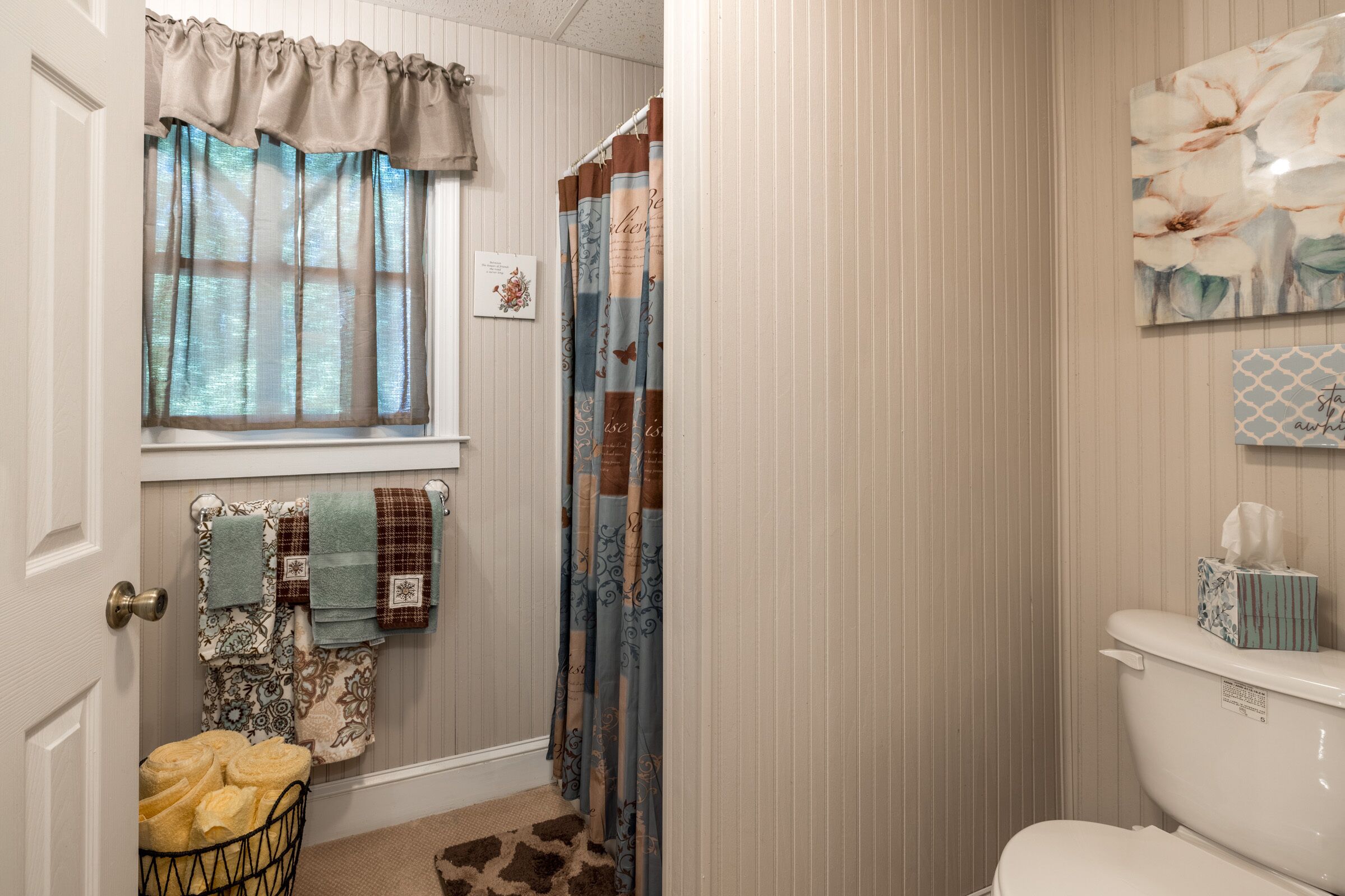 A bathroom with a toilet, a window with sheer curtains, a shower curtain, and a towel rack with multiple towels.