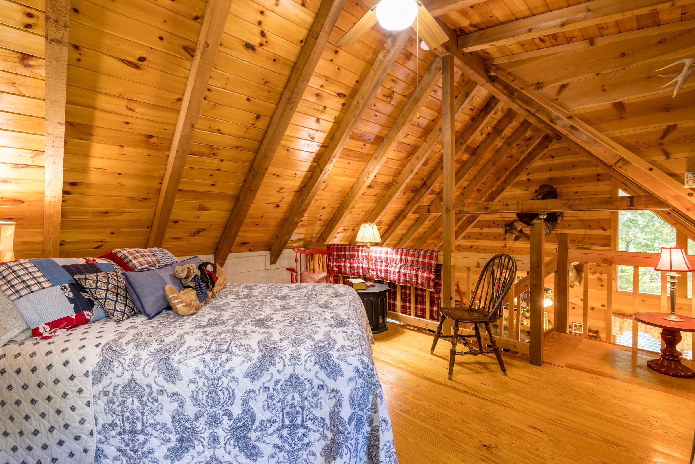 Un dormitorio acogedor en el ático con techo de madera, una cama con ropa de cama estampada, una silla y una mesita con lámpara.