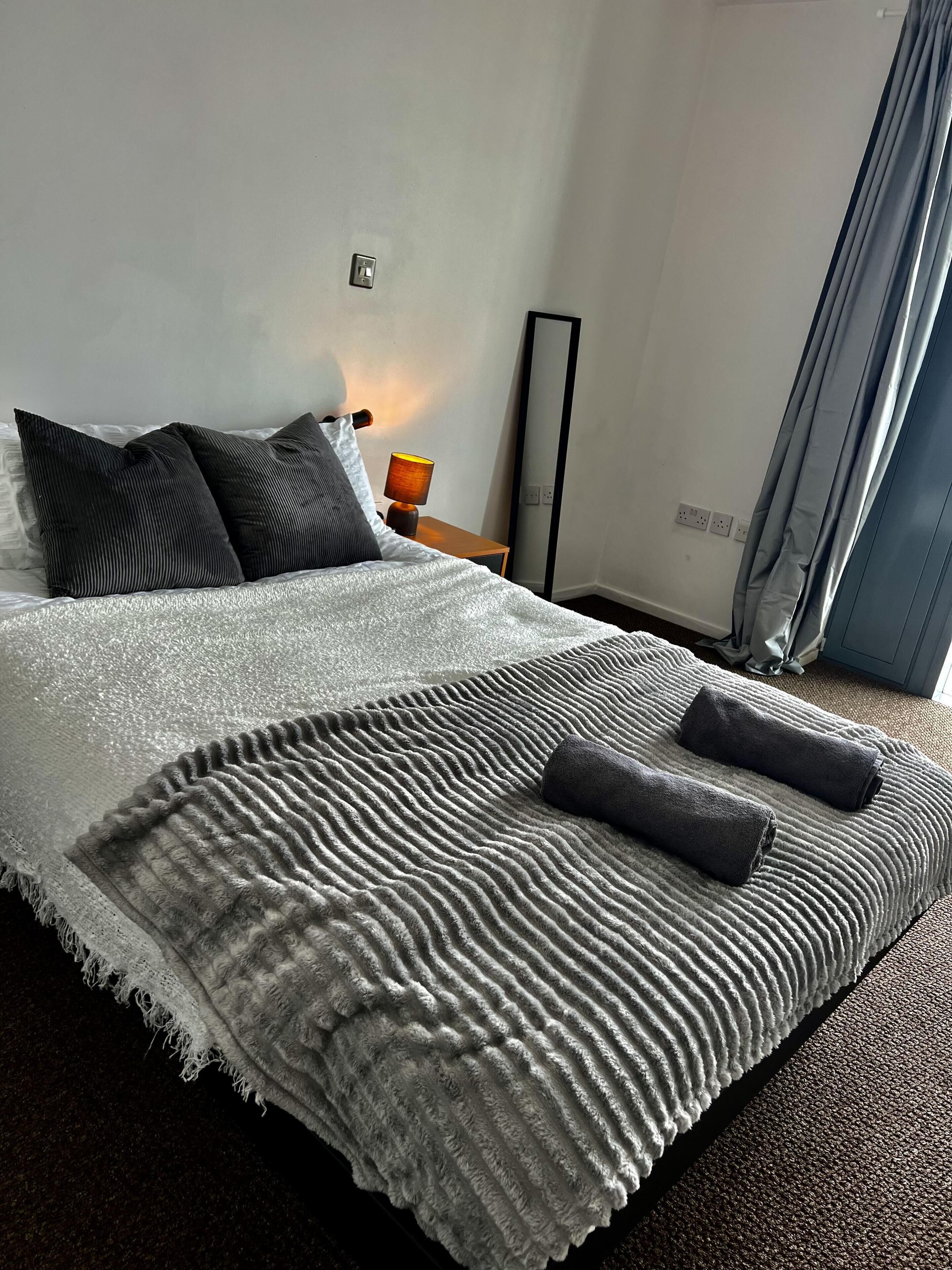 Un lit bien fait, recouvert d’une couverture blanche et grise à texture intéressante, avec deux oreillers foncés et deux serviettes pliées.