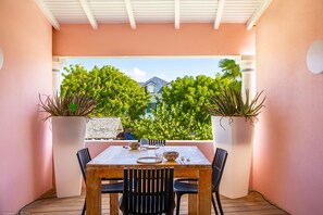 An outdoor patio with a wooden table set for dining, surrounded by chairs, and flanked by large potted plants.