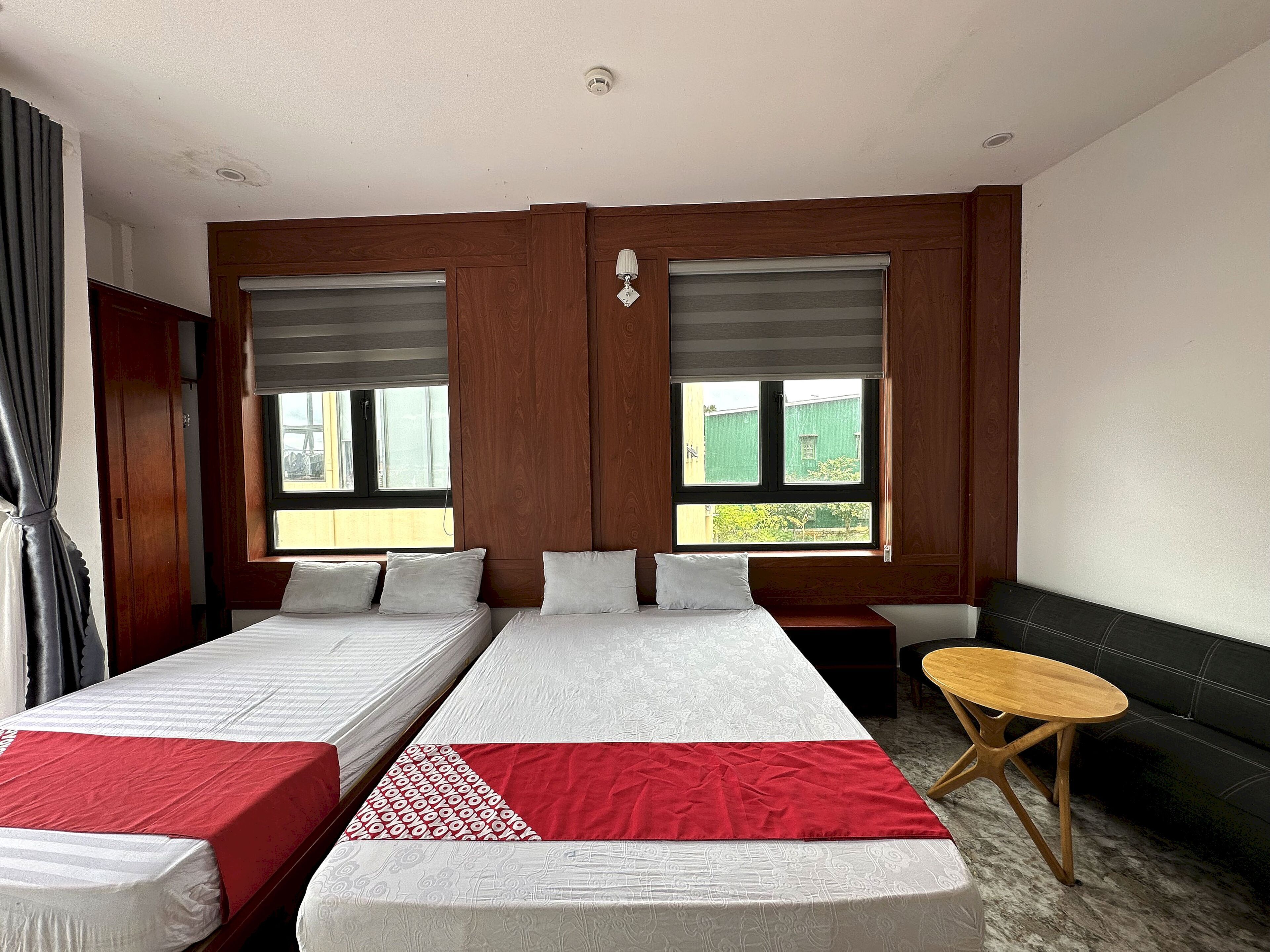 A hotel room with two single beds, a small wooden table, and a view of greenery through the windows.
