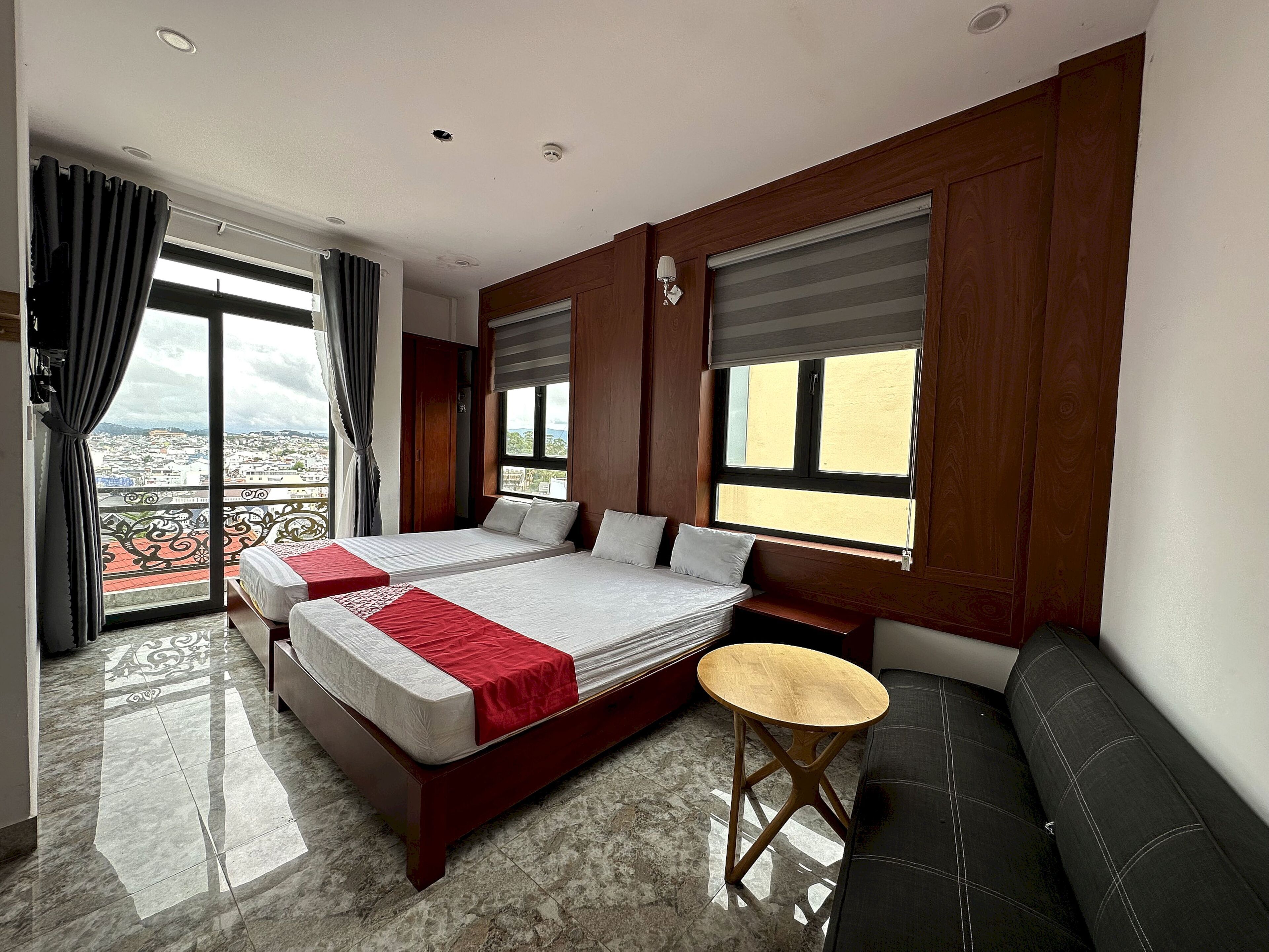 A hotel room with two beds, a small round table, a sofa, and large windows offering a city view.