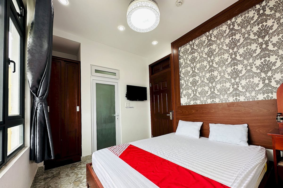 A hotel room with a bed, a wooden headboard, a patterned wallpaper, a red and white bedspread, a marble floor, a door, and a window with curtains.