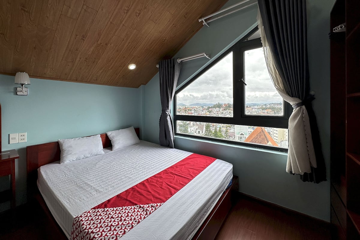 A bedroom with a large bed, a window with curtains, a wooden ceiling, and a view of a cityscape.