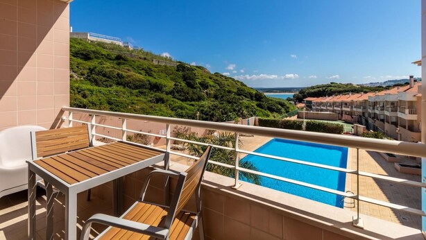 Un balcón con mesa y sillas de madera, una piscina y vistas a una colina y edificios.