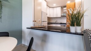 Una cocina moderna con armarios blancos, encimera negra y techo de madera. Hay una planta en el lado derecho y un jarro de vidrio en el izquierdo.