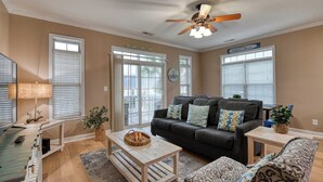 A living room with a dark grey sofa, a wooden coffee table, and a patterned ottoman. There is a ceiling fan, a lamp, and a window with blinds. - Spacious 4 Bedroom, 3.5 Bath Garden City Beach Home with Private Pool (Garden City Beach)