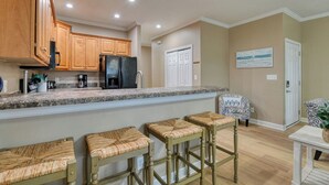 A kitchen with wooden cabinets, a granite countertop, and a black refrigerator. There are three wicker bar stools and a white door in the background. - Spacious 4 Bedroom, 3.5 Bath Garden City Beach Home with Private Pool (Garden City Beach)