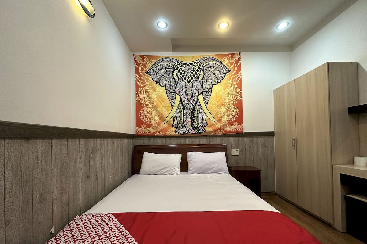 A hotel room with a bed, wooden headboard, a red and white patterned blanket, a wooden wardrobe, and a tapestry with an elephant design on the wall.