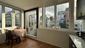 Quarto com mesa de jantar, cadeiras, cafeteira e janelas amplas com vista para edifícios.