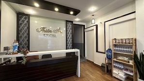 Uma área de recepção de hotel com design moderno, com um balcão de recepção, uma parede decorativa com uma figura religiosa e uma prateleira com água engarrafada e snacks.