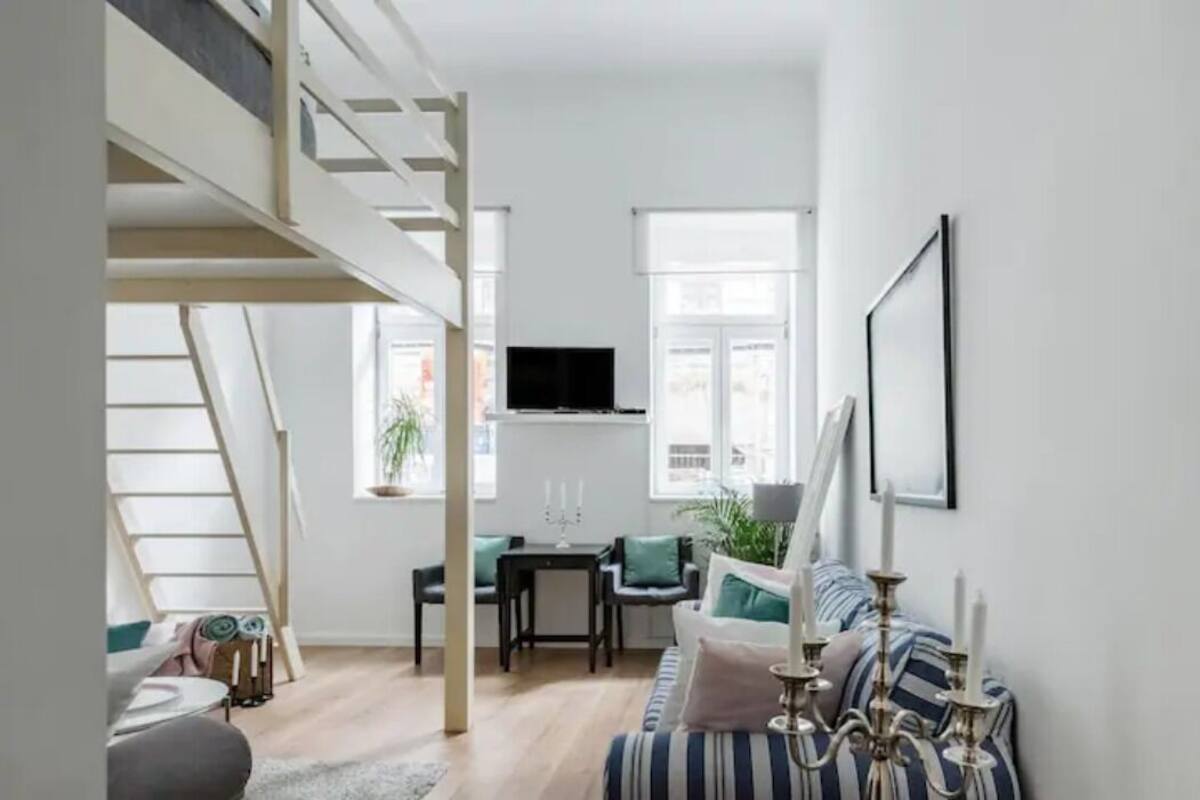 A modern living room with a loft bed, a flat-screen TV, a dining area, and a staircase.