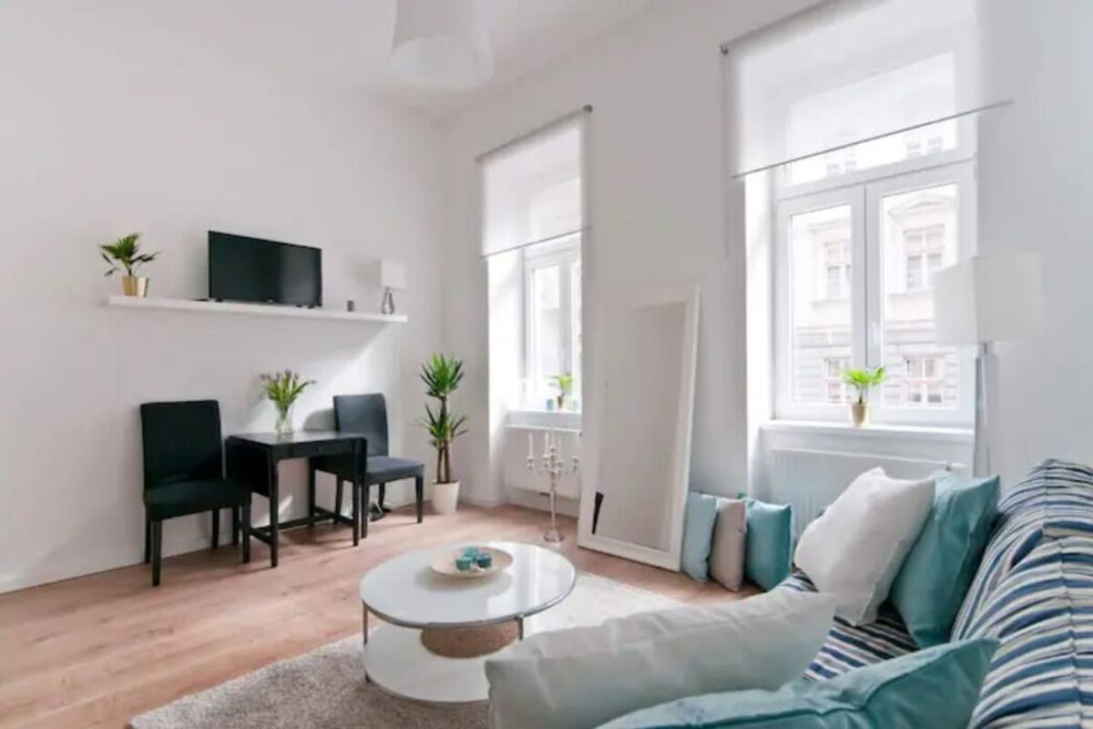 A modern living room with a sofa, a coffee table, and a TV on a shelf. There are plants and a window with blinds.