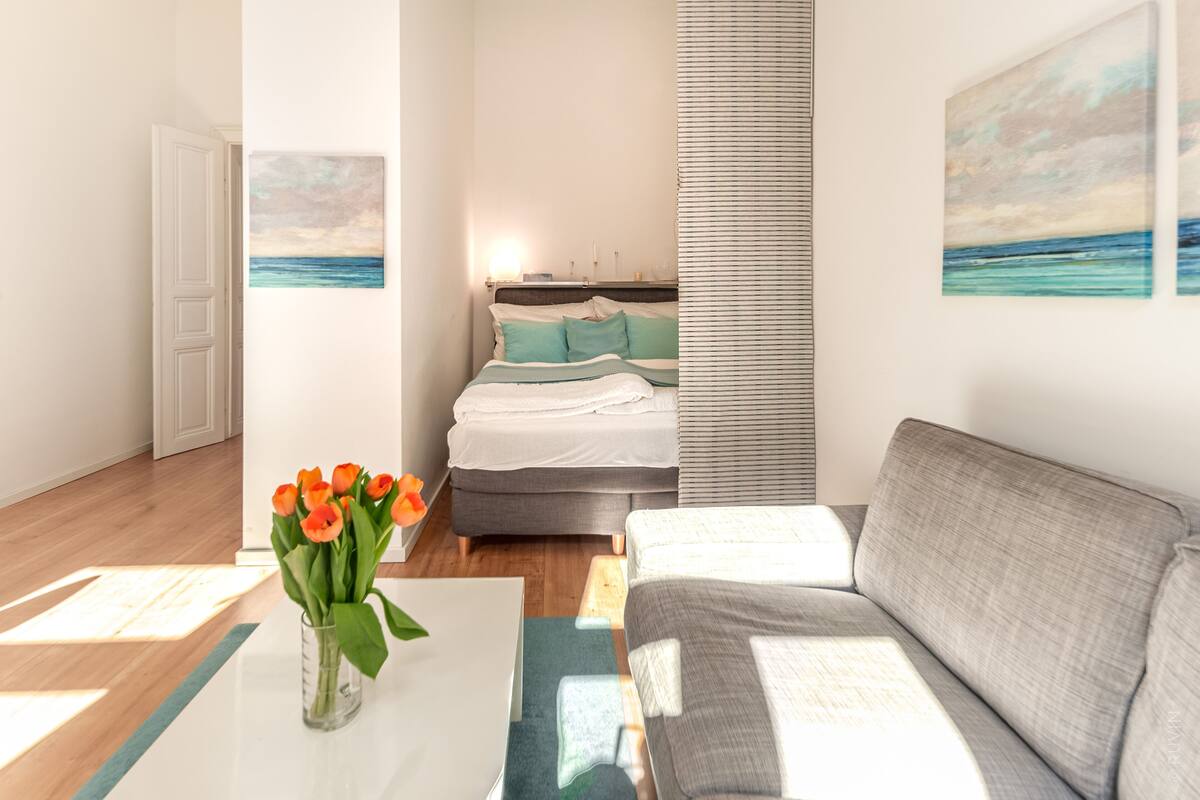 A bedroom with a bed, a grey sofa, a vase with flowers, and artwork on the walls.